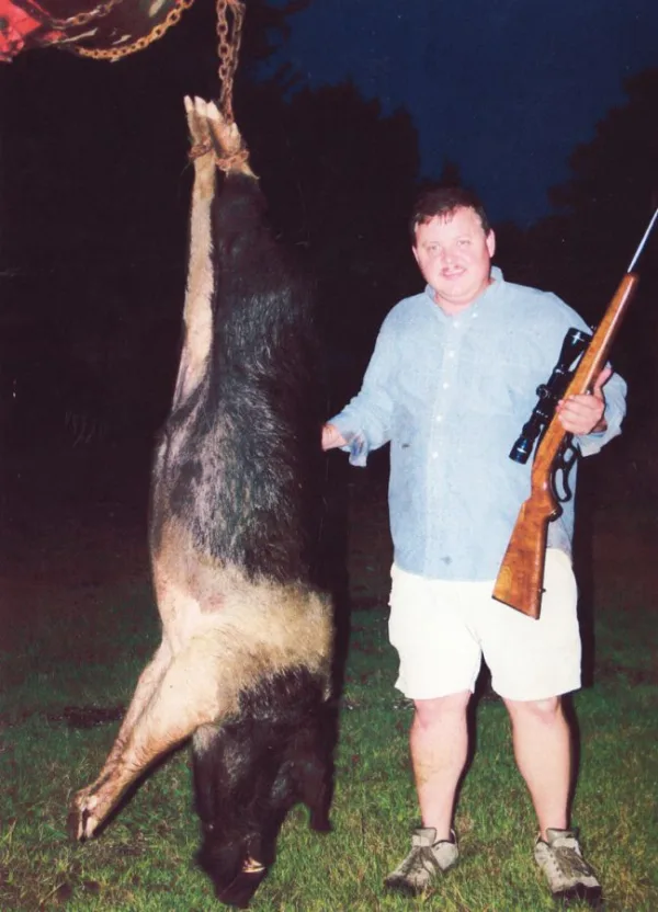 man with rifle next to dead hog strung up in a tree