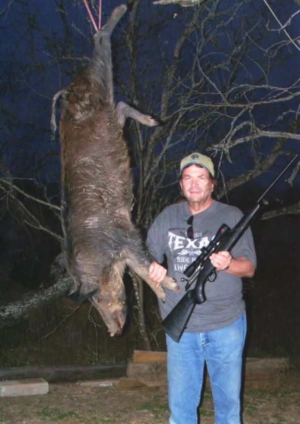 man with rifle next to dead hog strung up in a tree